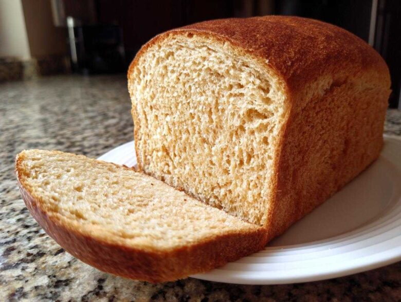 A freshly baked loaf of soft whole wheat bread with one thick slice cut and resting against the main loaf on a white plate.