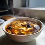 A close-up of a steaming white bowl filled with rich, dark hot and sour soup recipe, showing tofu and mushrooms.