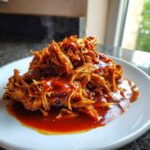 A close-up of steaming, shredded Slow Cooker Pulled Pork generously coated in rich BBQ sauce on a white plate.