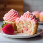 A halved strawberry cupcake showing the moist interior with strawberry pieces, next to a whole fresh strawberry and another frosted cupcake.