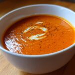 Close-up of a white bowl filled with vibrant orange tomato bisque, garnished with a swirl of cream and cracked black pepper.