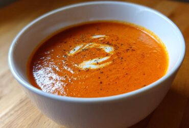 Close-up of a white bowl filled with vibrant orange tomato bisque, garnished with a swirl of cream and cracked black pepper.