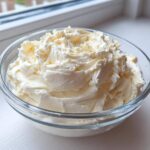 A clear glass bowl filled with freshly whipped, creamy vanilla buttercream frosting sitting on a white surface.