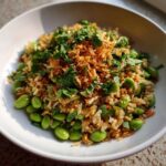 A white bowl filled with crispy rice salad mixed with bright green edamame and topped with fresh herbs and crispy fried onions.