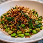 A vibrant serving of crispy rice salad featuring bright green edamame, fresh mint, and a generous topping of crispy fried rice.