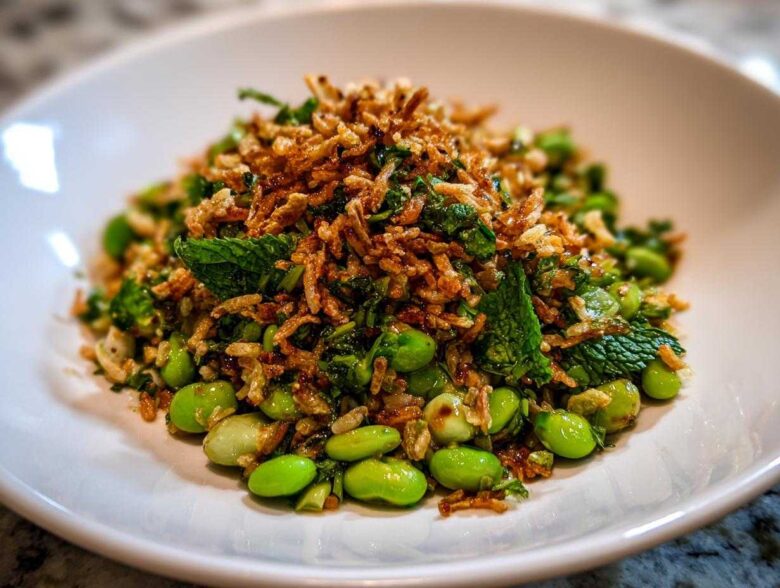 A vibrant serving of crispy rice salad featuring bright green edamame, fresh mint, and a generous topping of crispy fried rice.