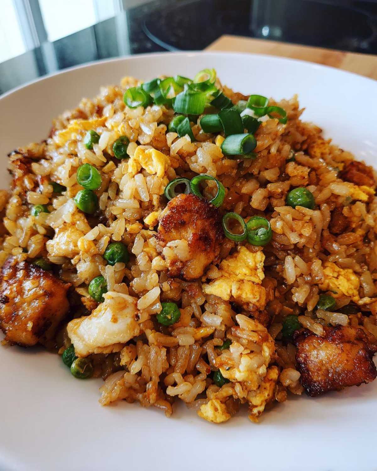 A close-up of a white bowl filled with flavorful chicken fried rice, mixed with scrambled egg, peas, and topped with fresh green onions.