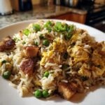 A close-up of a serving of homemade chicken fried rice with visible chunks of browned chicken, scrambled egg, and green peas, topped with scallions.