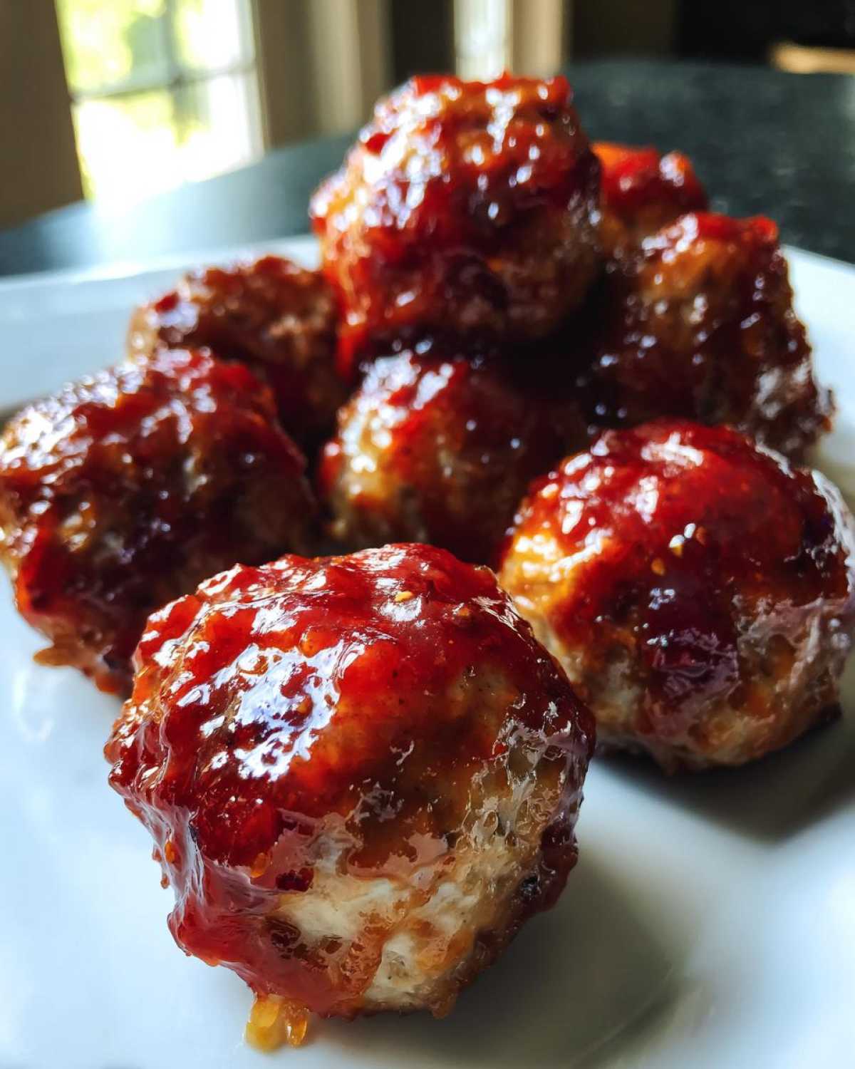Close-up of several glossy cranberry meatballs coated in a thick, sweet-and-sour red glaze on a white plate.