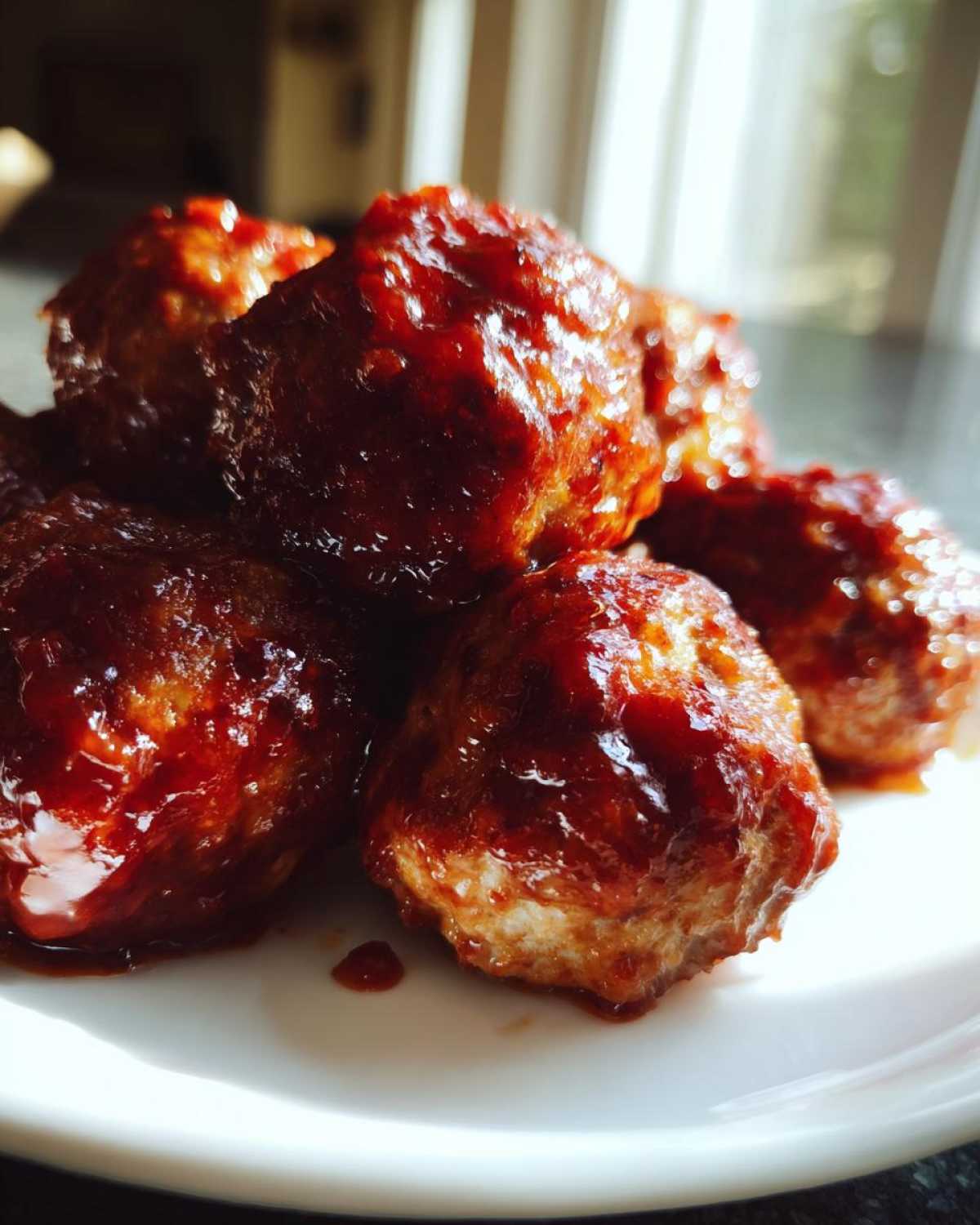 A close-up shot of several glistening cranberry meatballs piled on a white plate, coated in a rich, dark red sauce.