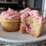 Two strawberry cupcakes on a white plate, one whole and one bitten to show the pink filling inside the cake base.