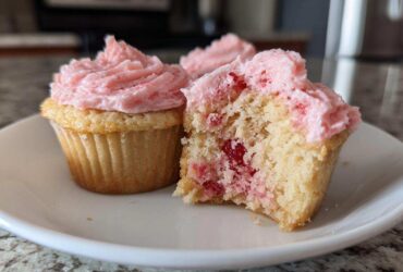 Two strawberry cupcakes on a white plate, one whole and one bitten to show the pink filling inside the cake base.