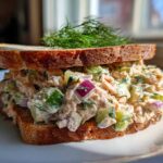 A thick sandwich filled with creamy salmon salad, diced cucumber, and red onion, topped with fresh dill.