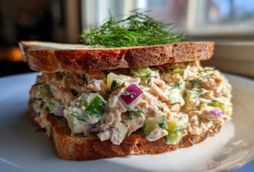 A thick sandwich filled with creamy salmon salad, diced cucumber, and red onion, topped with fresh dill.