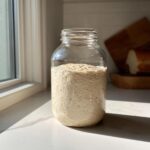 A highly active sourdough starter, full of bubbles, filling a large glass jar placed on a sunny kitchen counter.