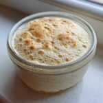A jar containing a very active sourdough starter, showing a thick layer of foam and bubbles on top.