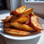A close-up of a pile of golden brown, crispy air fryer potato wedges seasoned with spices on a white plate.