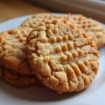 Close-up of several golden-brown peanut butter cookies with classic crosshatch fork marks stacked on a white plate.