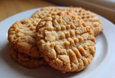 Close-up of several golden-brown peanut butter cookies with classic crosshatch fork marks stacked on a white plate.