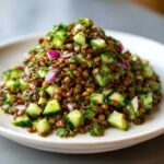 A mound of vibrant lentil salad mixed with diced cucumbers, red onion, and fresh herbs on a light plate.