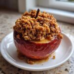 A single, glistening baked apple topped high with brown sugar crumble, served on a white plate.