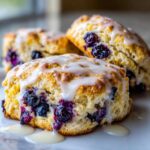 Close-up of a tall, flaky blueberry scone topped with dripping vanilla glaze.