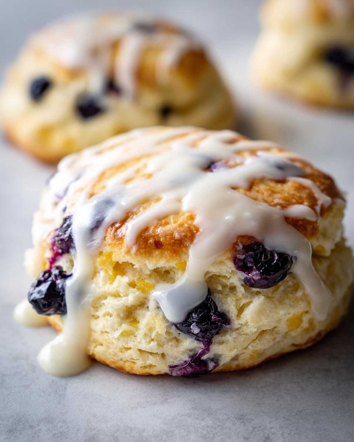 Close-up of a tall, flaky blueberry scone topped with a thick vanilla glaze dripping down the sides.