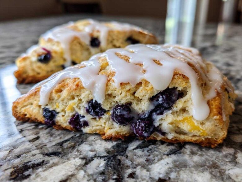 Close-up of a freshly baked blueberry scone, drizzled with white glaze, showing a flaky texture and burst blueberries.