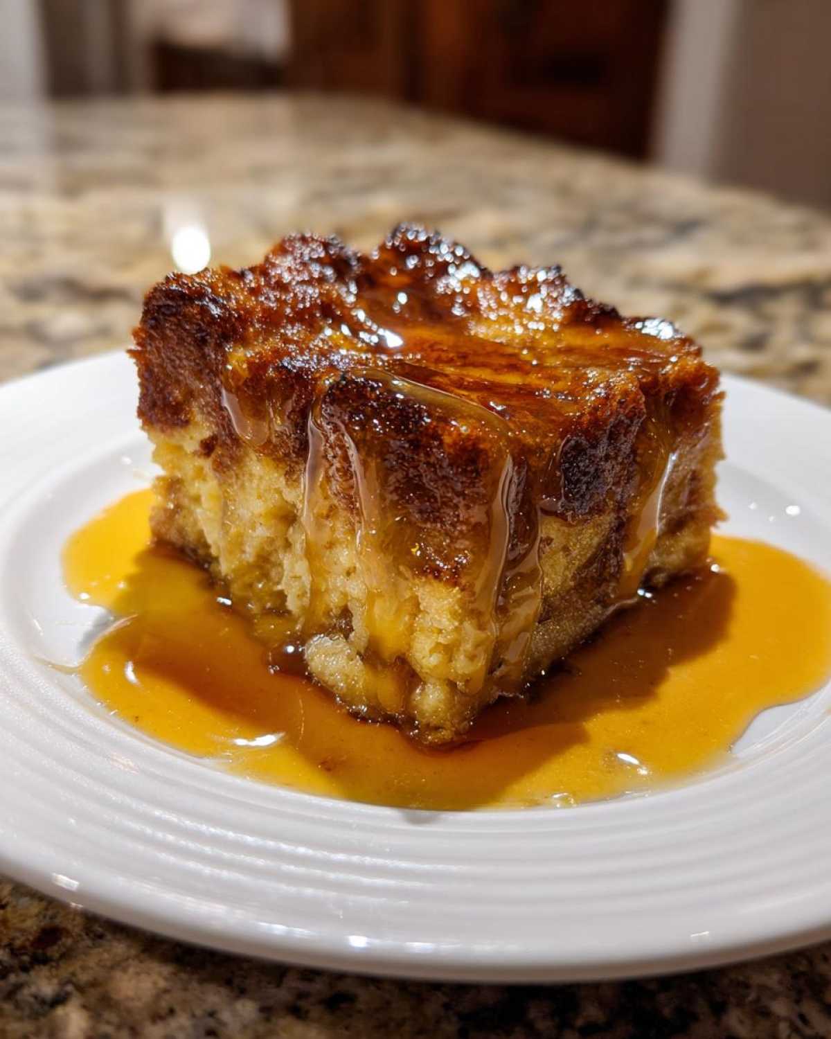 A square serving of rich, caramelized bread pudding dripping with a thick, golden sauce on a white plate.