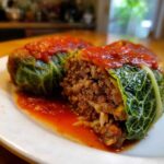 Close-up of two cabbage rolls, one cut open revealing the ground meat and rice filling, topped with red tomato sauce.