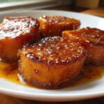 Four thick slices of glistening candied sweet potatoes covered in a rich, amber glaze served on a white plate.