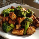 Close-up of a serving of glossy, saucy chicken and broccoli stir-fry on a square white plate.
