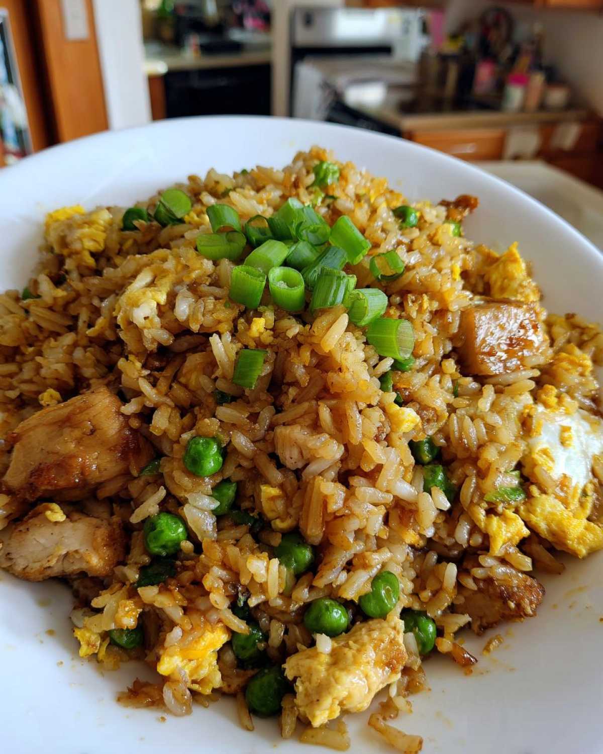 A close-up of a white bowl filled with savory chicken fried rice, mixed with peas and egg, topped with fresh green onions.