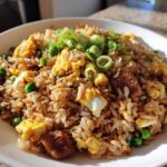 A close-up of a white bowl filled with flavorful chicken fried rice, featuring scrambled egg, peas, and green onion garnish.