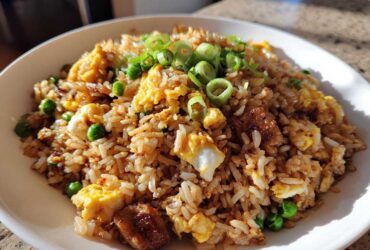 A close-up of a white bowl filled with flavorful chicken fried rice, featuring scrambled egg, peas, and green onion garnish.