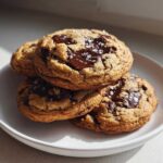 A stack of four gooey chocolate chip cookies, topped with melted dark chocolate and flaky sea salt.