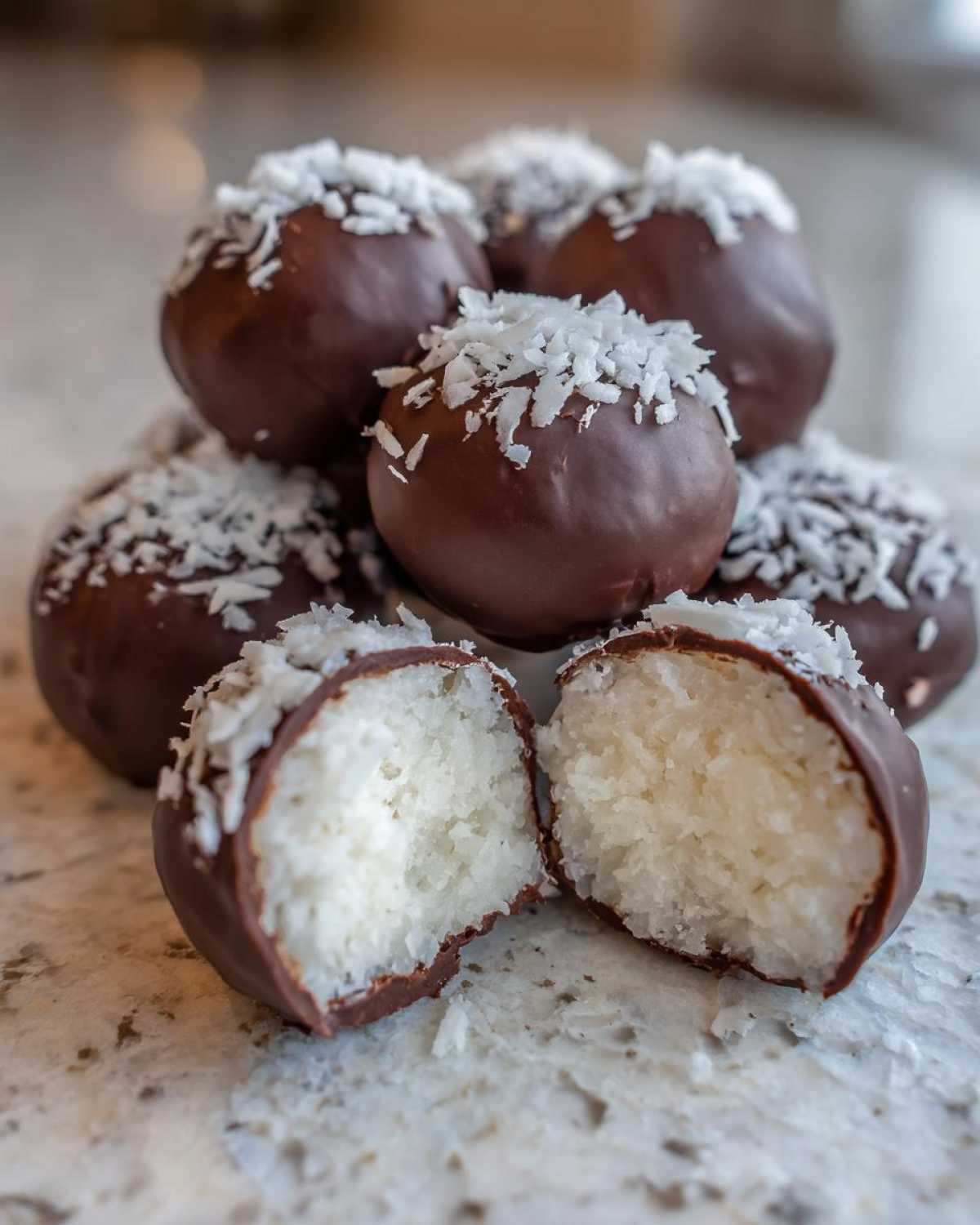 A stack of chocolate covered coconut balls, one cut in half revealing the white coconut filling, topped with shredded coconut.