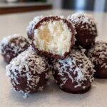 A pile of homemade coconut balls dipped in dark chocolate and rolled in shredded coconut, one is cut open showing the white interior.