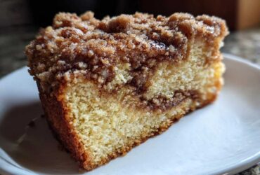 A close-up shot of a moist slice of coffee cake featuring a thick cinnamon swirl and crumb topping on a white plate.