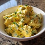 Close-up of creamy, yellow egg salad recipe garnished with chopped green onions in a white bowl.