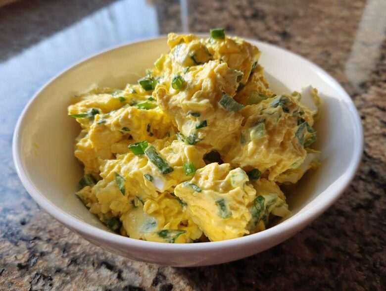 Close-up of creamy, yellow egg salad recipe garnished with chopped green onions in a white bowl.