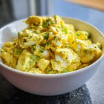 Close-up of chunky, creamy yellow egg salad recipe with visible pieces of celery and fresh herbs in a white bowl.