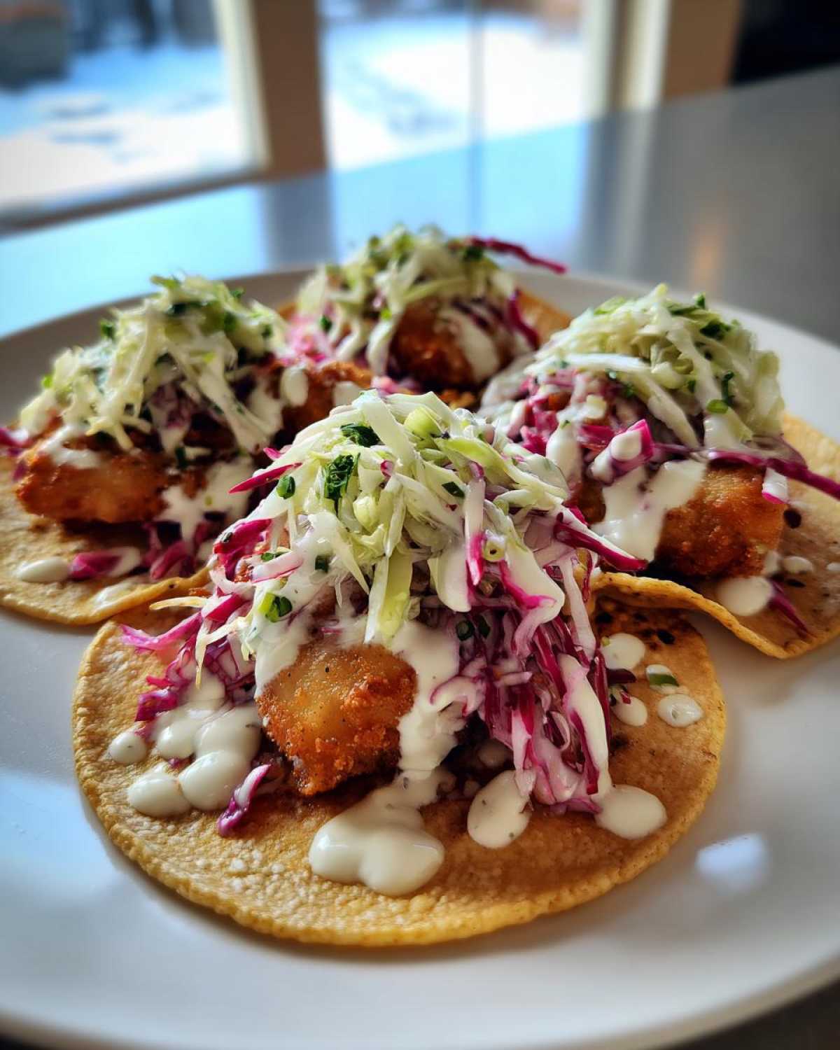 Four golden, crispy fish tacos topped with red cabbage slaw and a drizzle of white sauce, ready to eat.