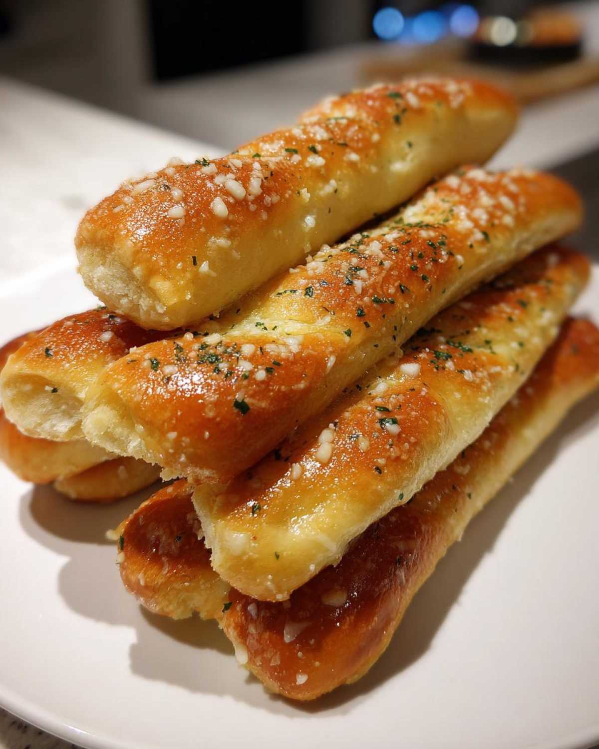 A close-up stack of golden brown, glazed breadsticks topped with minced garlic and parsley.
