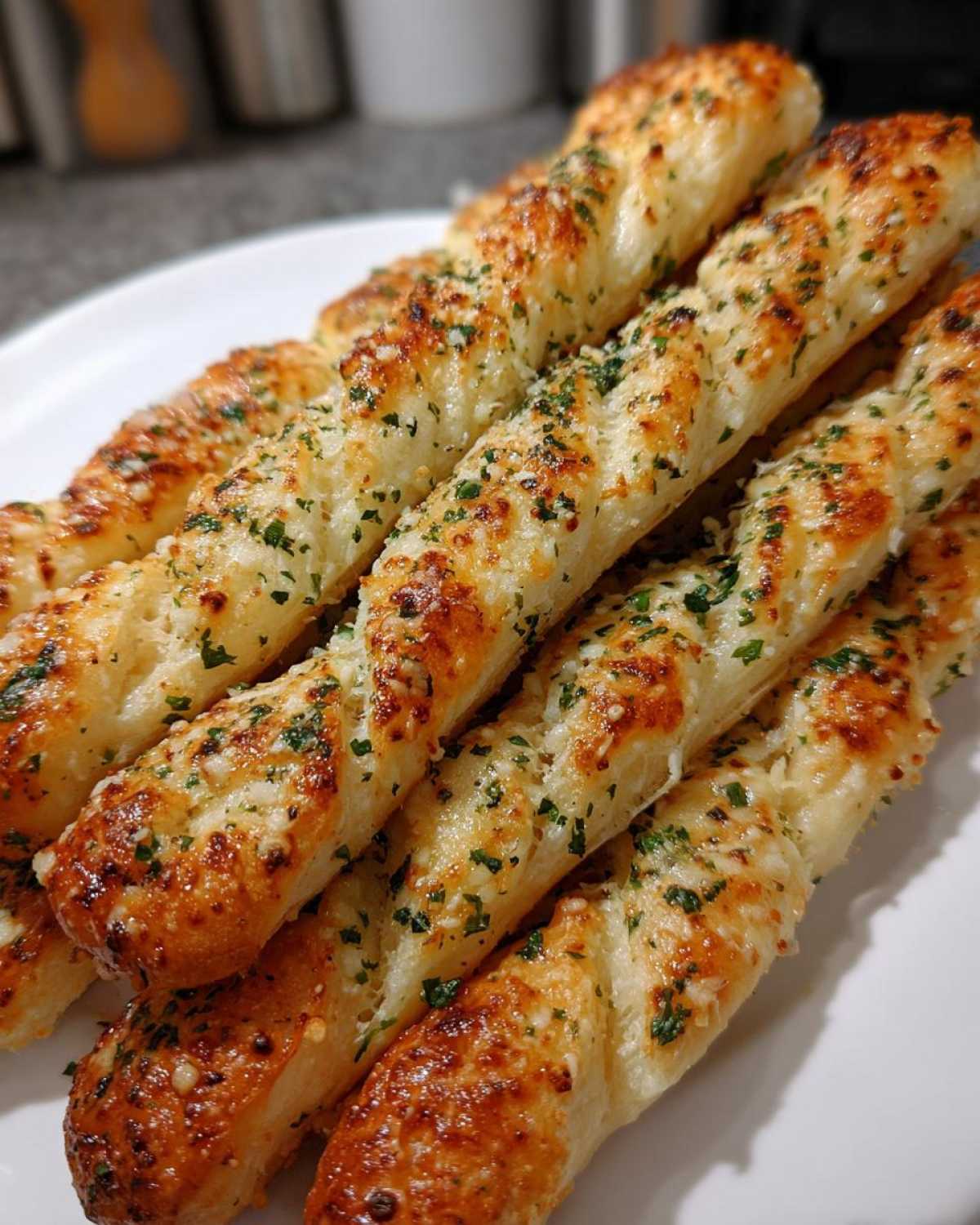 A close-up of several golden brown, cheesy garlic parmesan breadsticks stacked on a white plate.