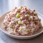 A close-up of a generous mound of creamy ham salad mixed with celery and bacon bits, served on a white plate.