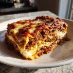 A thick, layered slice of rich meat lasagna with bubbly cheese topping, served on a white plate.