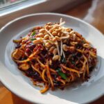 A close-up of a serving of rich, dark sauced Pad Thai noodles topped with roasted peanuts and fresh bean sprouts.