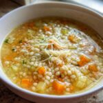 Close-up of a white bowl filled with amazing pastina soup, featuring small pasta, carrots, celery, and shredded cheese.