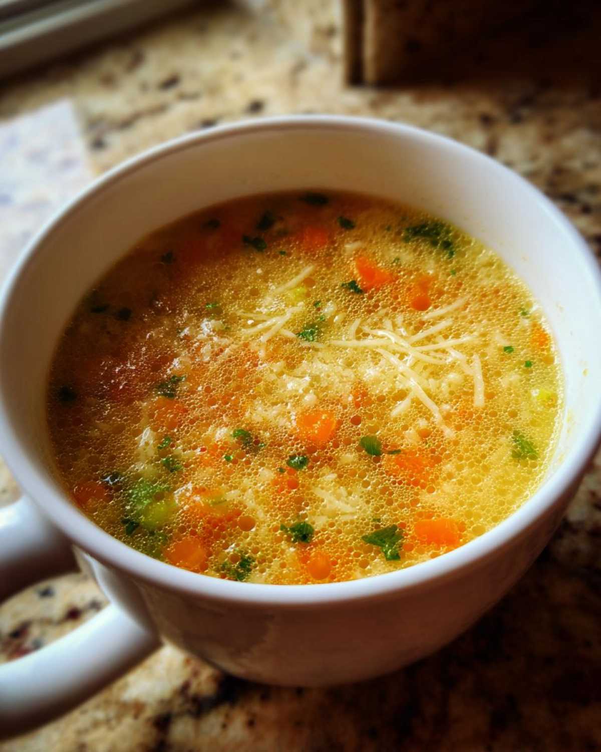 Close-up of a white mug filled with hot pastina soup, featuring broth, tiny pasta, carrots, and herbs.